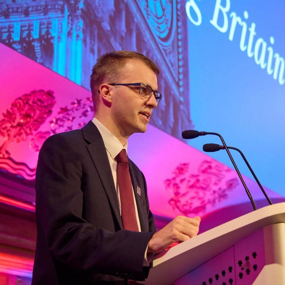 Adam Williams speaking at an SDP conference podium, with a projection screen reading '...s Britain' behind him